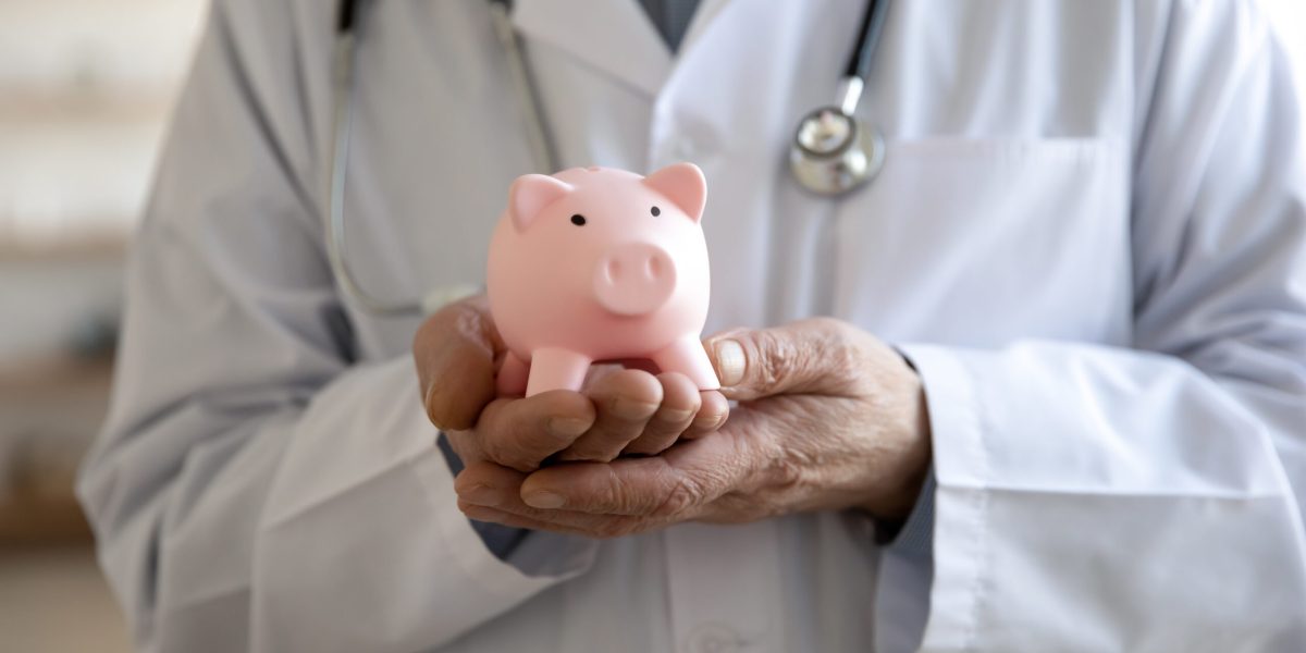 Close up of elderly male doctor in white medical uniform hold piggybank for hospital charity fee or contribution. Mature man therapist ask for volunteer donation in clinic budget. Welfare concept.